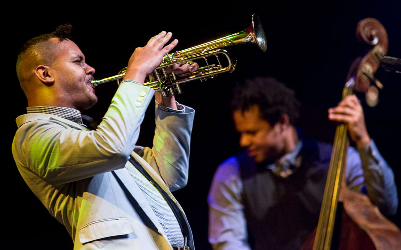 Ahamefule Oluo dressed in a suit, playing a trumpet and an out of focus Evan Flory-Barnes playing bass in the background.