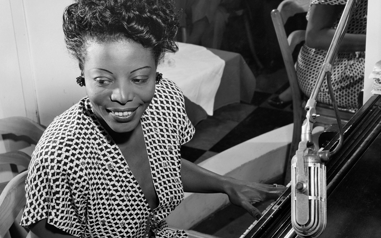 Mary Lou Williams seated at the piano, playing and smiling.