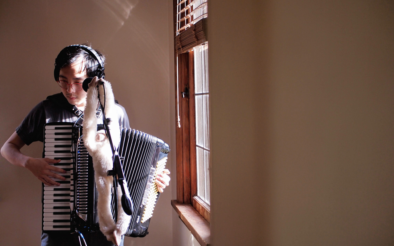 Josh Hou playing an accoordion with a window to the right of him.