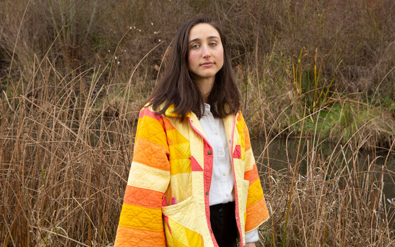 Abbey Blackwell wearing a colorful patchwork jacket, standing in front of wild grass.