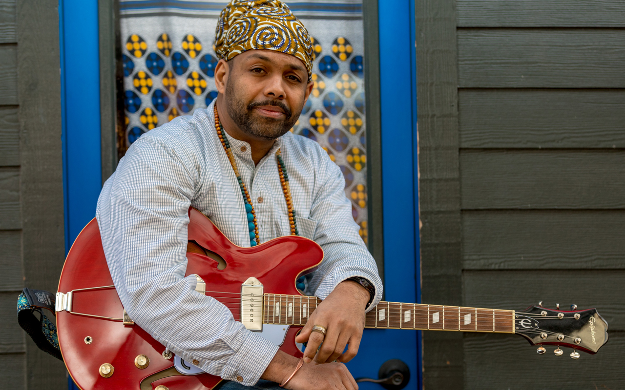 Benjamin Hunter with a red guitar, seated in front of a blue door.