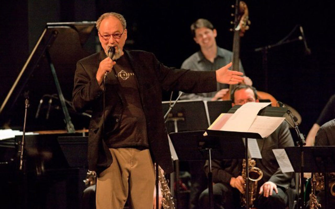 Jim Knapp with microphone in hande, speaking to an audience, with musicians in the background.