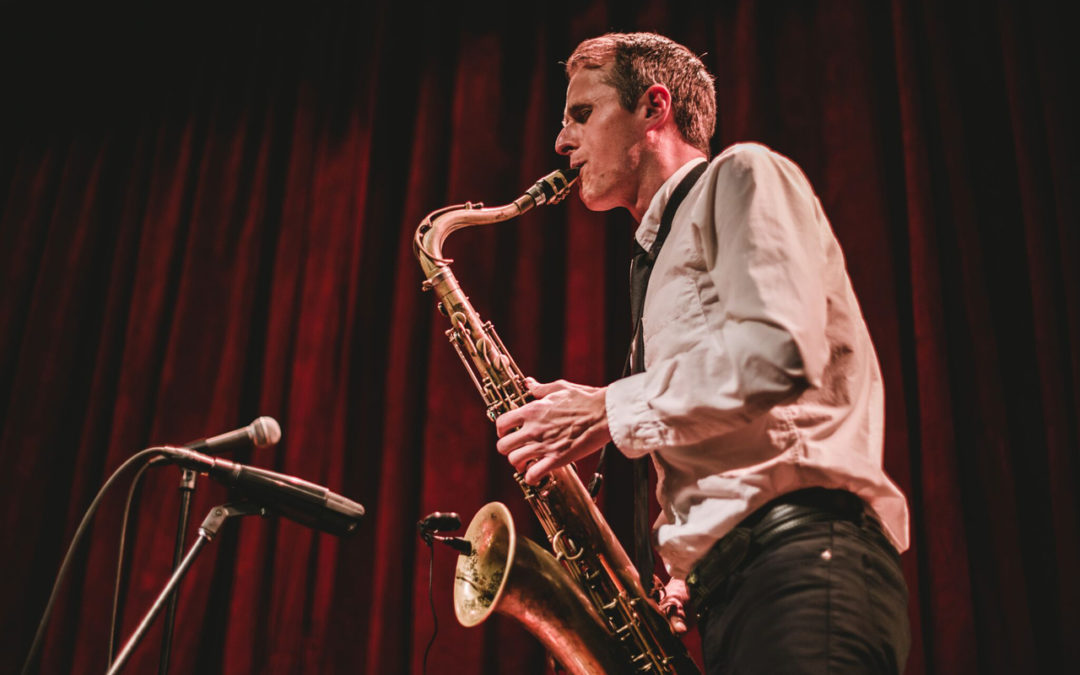 Neil Welck playing a saxophone in profile with a red curtain behind him.