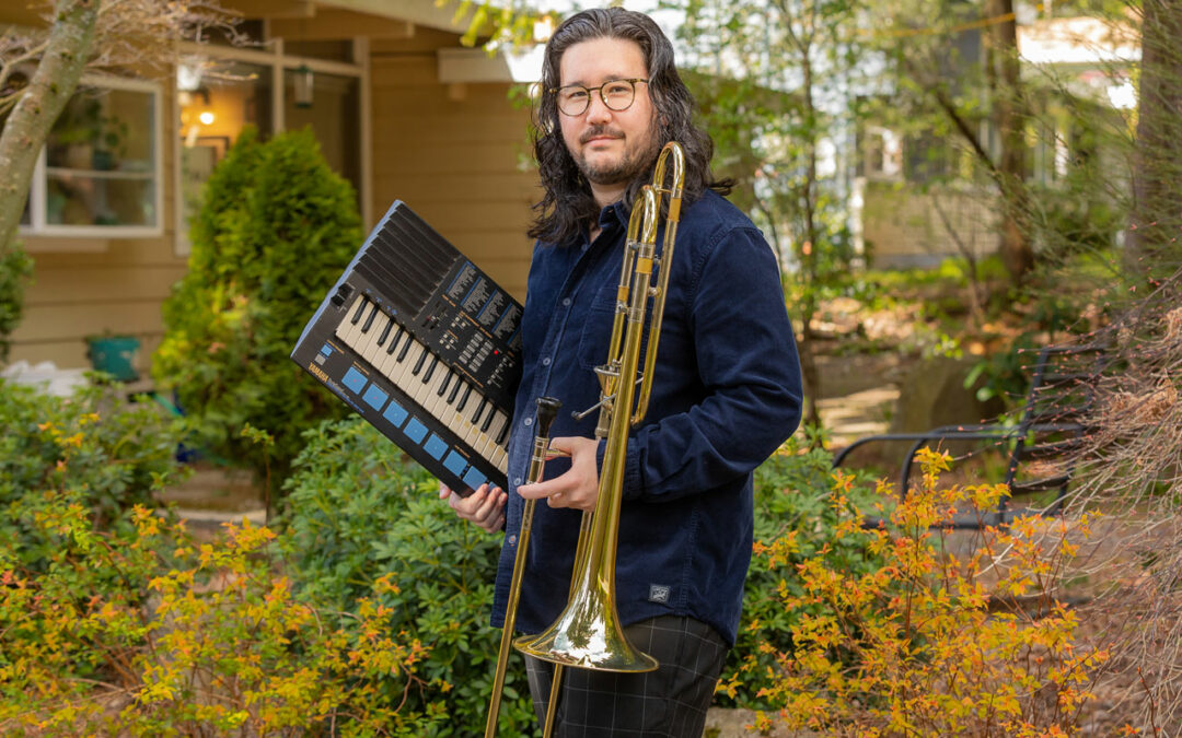 Greg Kramer holding a trombone and a keyboard standing in a yard filled with trees of various colors.