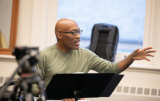 Darrell Grant talking by a music stand with arm outstretched wearing a green long-sleeved t-shirt.