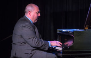 Eric Velinde wearing a suit, playing the piano in profile.