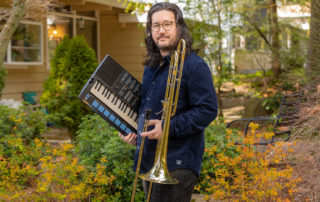 Greg Kramer holding a trombone and a keyboard standing in a yard filled with trees of various colors.
