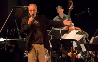 Jim Knapp with microphone in hande, speaking to an audience, with musicians in the background.