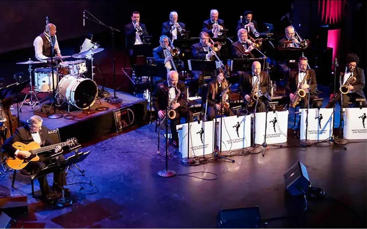 Seattle Repertory Jazz Orchestra on stage. From left to right with guitar and drums on left and 3 rows of artists on the right. Trumpets on top row, trombones in middle, and saxophones in the first row with traditional "front" music stands with white background and black SRJO logo.